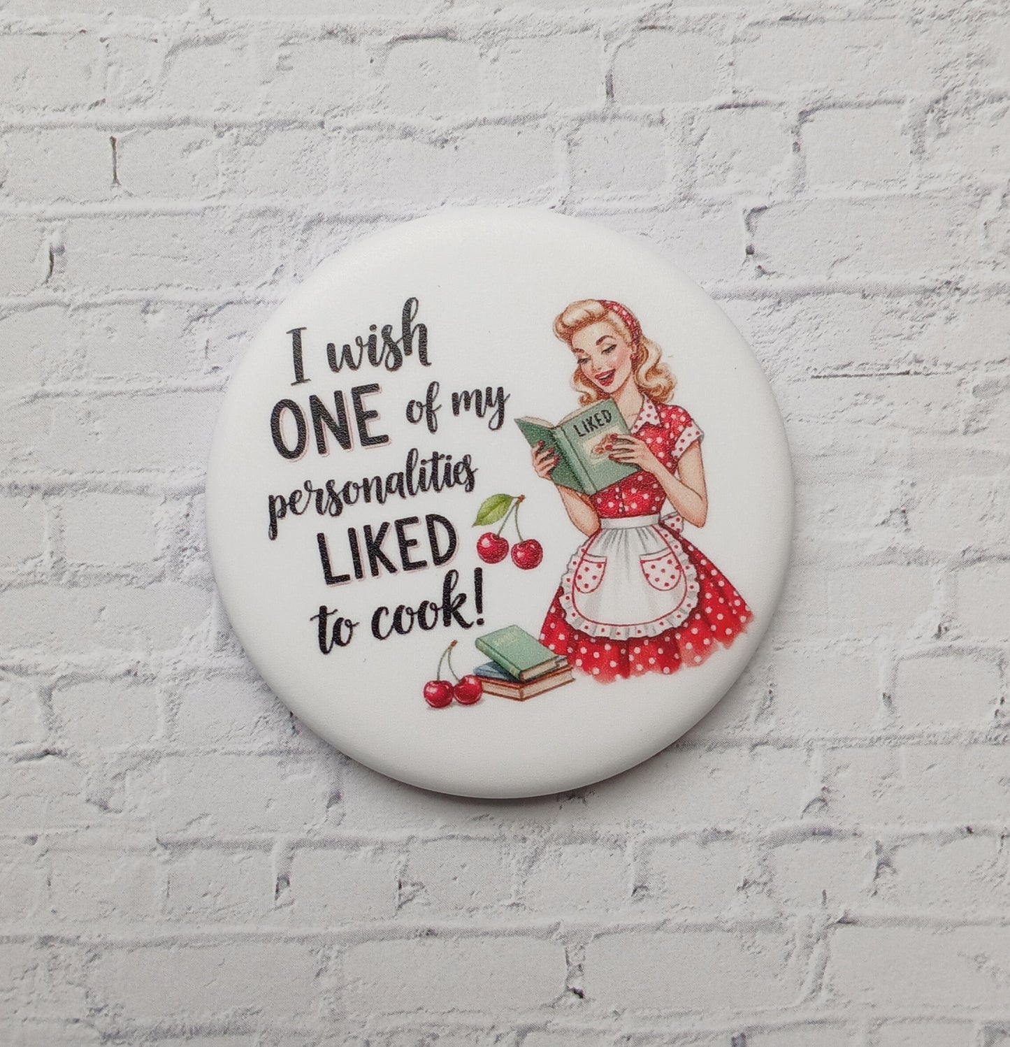 A white, round refrigerator magnet with a white brick background. There is a retro woman with a cookbook. The magnet reads I wish one of my personalities loved to cook!